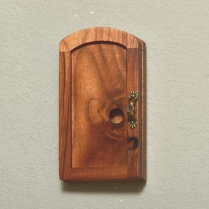 May include: A small, wooden cabinet with a curved top and a brass handle. The cabinet is made of a rich, dark wood with visible grain patterns. A small, round hole is visible on the door, and a crescent moon is carved into the door.