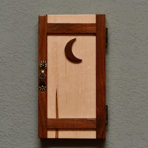 May include: A small wooden door with a crescent moon cutout. The door is framed in dark brown wood with a light wood panel and a decorative brass handle. The door is hinged and mounted on a gray wall.