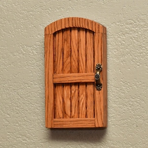 May include: A wooden miniature door with a curved top and vertical planks. It features a horizontal bar and an ornate bronze-colored handle. The door is mounted on a textured, light-colored wall.