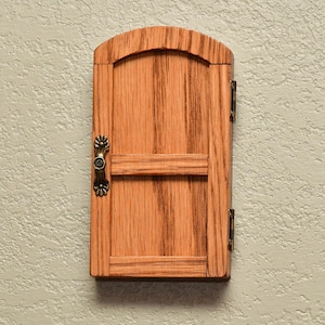 May include: A small, arched wooden door with a natural light brown wood grain finish. The door features a horizontal crossbar, a decorative antique brass-colored metal handle on the left side, and two small metal hinges attached to the right side.
