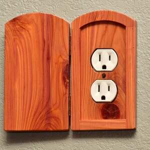 May include: A reddish-brown wooden electrical outlet cover with a hinged door. The cover features a natural wood grain pattern and an arched top. The door is open, revealing a standard white dual electrical outlet mounted within the wooden frame.