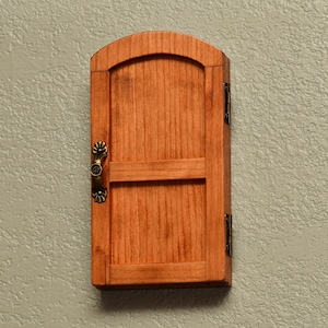 May include: A small, wooden cabinet with a door. The cabinet is stained a warm reddish-brown color and has a decorative handle and hinges. The door has a rounded top and a horizontal bar across the middle.