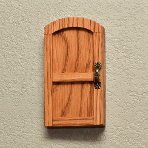 May include: A small, arched wooden door with a natural light brown wood grain finish. The door features a horizontal crossbar and a decorative, antique-style brass-colored metal handle attached to the right side, providing a detailed accent.