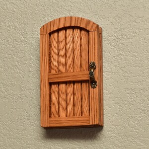May include: A small, wooden door with an arched top and vertical slats. The door is made of light brown wood and has a horizontal bar across the middle. A decorative, bronze-colored handle is on the right side. The door is mounted on a textured, light-colored wall.