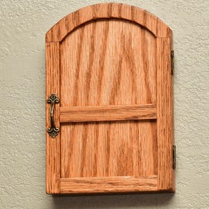 May include: A rectangular wooden cabinet door with an arched top, featuring a natural light brown wood grain finish. The door includes a horizontal crossbar, a decorative antique brass-colored metal handle on the left, and two brass hinges on the right.