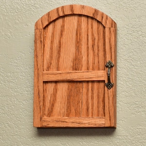 May include: A small, wooden door with an arched top and a decorative handle. The door is made of reddish-brown wood with a visible grain pattern. The handle is dark metal, and the door is mounted on a light green wall.