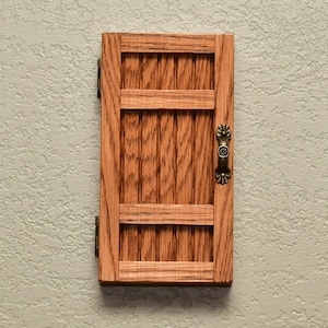 May include: A rectangular wooden door with a natural light brown wood grain finish, featuring vertical panels, two horizontal crossbars, two metal hinges on the left side, and an ornate, antique-style brass handle on the right side.