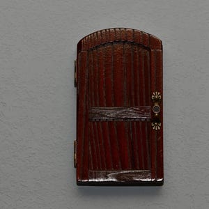 May include: A small wooden door with a brown stain and a decorative metal handle. The door has a raised arch and a horizontal panel in the center.