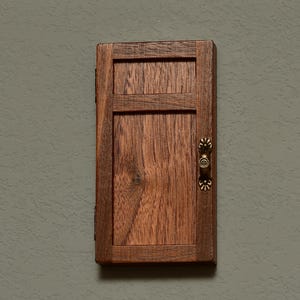 May include: A small, rectangular wooden cabinet with a dark brown finish. The cabinet door has a recessed panel design and a decorative brass handle. The wood grain is visible, adding texture to the piece. The cabinet is mounted on a gray wall.