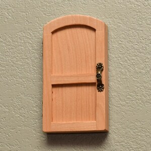 May include: A small, arched wooden door made of light-colored, natural wood. The door features a horizontal crossbar and a decorative, antique-style brass handle attached to the right side. The wood grain is visible across the flat surface.