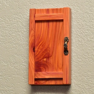 May include: A rectangular wall-mounted wooden cabinet with a single door featuring a prominent reddish-orange wood grain pattern. The cabinet is equipped with a decorative antique bronze-colored metal handle attached to the right side of the door.