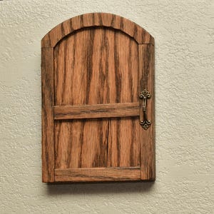 May include: A small, wooden arched door with a dark brown finish and a decorative brass handle. The door has a horizontal brace and vertical wood grain. The door is mounted on a light beige wall.