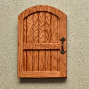 May include: A small, wooden fairy door with an arched top and a decorative handle. The door is made of vertical wooden planks and has a horizontal crossbar. The door is stained a warm, natural wood color and is mounted on a textured wall.