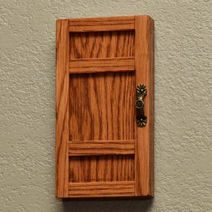 May include: A rectangular wooden wall-mounted cabinet door featuring a medium brown oak wood grain finish. The door has a vertical panel design with two horizontal crossbars and a decorative antique brass-toned metal handle on the right side.
