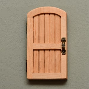 May include: A small, wooden door with a rounded top and vertical slats. The door has a decorative bronze handle and hinges, and is mounted on a textured, gray wall. The door is made of light-colored wood.