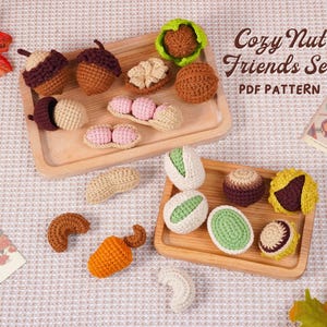 May include: A collection of handmade crochet nuts and seeds, including acorns, peanuts, and pistachios, arranged on wooden trays. The image includes the text "Cozy Nut Friends Set PDF Pattern". The nuts are in various colors, such as brown, beige, green, and orange.