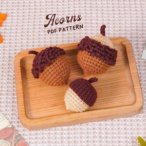 May include: Three crocheted acorns in shades of brown and beige sit on a wooden tray. The acorns have a textured appearance, with a dark brown cap and a lighter brown or beige body. The text "Acorns PDF PATTERN" is visible.