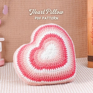 May include: A heart-shaped crochet pillow in shades of pink and white. The pillow features concentric circles of color, creating a gradient effect. The text "Heart Pillow PDF Pattern" is visible at the top. The pillow is on a white surface.