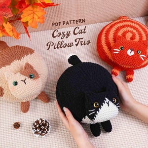 May include: Three handmade cat-shaped pillows in cream, black, and orange. The pillows have embroidered facial features and are arranged on a textured surface. The text "Cozy Cat Pillow Trio" is visible.