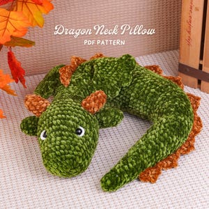 May include: A green and brown crocheted dragon neck pillow with black and white eyes. The text Dragon Neck Pillow PDF PATTERN is visible above the plush toy. The dragon has a textured yarn finish, small horns, and a long, curved body shape.