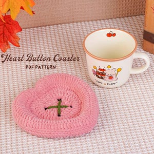 May include: A pink, heart-shaped crochet coaster with a green cross stitch detail, next to a cream-colored mug with a cartoon design. The mug features a cake and a pig illustration. The text "Heart Button Coaster PDF Pattern" is visible.