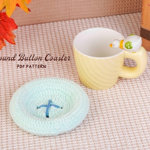 May include: A light yellow mug with a duck-shaped handle sits next to a light blue crocheted coaster with a blue cross-stitch design. The text "Round Button Coaster PDF Pattern" is visible. Autumn leaves are in the background.
