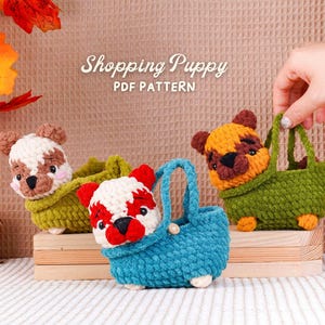 Op de afbeelding: Drie gehaakte puppyhondenspeeltjes in groene en blauwe manden, en één in een groen handmandje. De puppy's hebben bruine, witte en rode markeringen. De tekst "Shopping Puppy PDF PATTERN" staat boven de speeltjes.