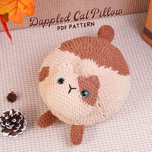 May include: A cream and brown, round cat pillow with green eyes and a black stitched nose and mouth. The pillow has the text "Dappled Cat Pillow PDF PATTERN".