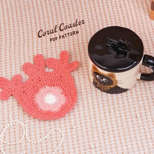 May include: A coral-colored crochet coaster shaped like a piece of coral with a white center, positioned next to a ceramic mug with a black lid and a cat illustration. The text Coral Coaster PDF PATTERN is visible in the upper left corner.