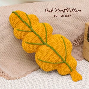 Könnte beinhalten: Ein handgefertigtes, gelbes Eichenblattkissen mit grünen Aderdetails. Das Kissen besteht aus einem strukturierten Material und liegt auf einer neutralfarbenen Decke. Der Text auf dem Bild lautet "Oak Leaf Pillow PDF Pattern".