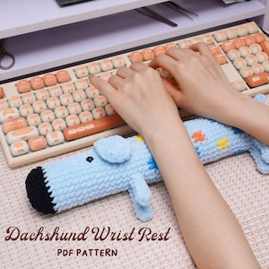 Peut inclure: Un repose-poignet en forme de teckel bleu clair, crocheté, avec un nez noir et de petites pattes bleues. Le teckel a de petits motifs floraux colorés. Le texte "Dachshund Wrist Rest PDF Pattern" est en bas de l'image.