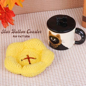 May include: A yellow star-shaped coaster with a red cross design, next to a white mug with a black lid and handle. The mug features a cat illustration. The text "Star Button Coaster PDF Pattern" is visible.