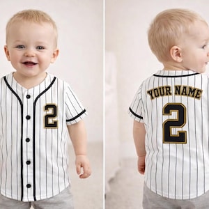 Könnte beinhalten: Ein weiß-schwarz gestreiftes Baseball-Trikot mit der Nummer 2 in Gold und Schwarz auf der Vorder- und Rückseite. Auf der Rückseite des Trikots steht außerdem der Text "YOUR NAME" in Gold. Das Trikot hat schwarze Verzierungen und Knöpfe.