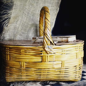 May include: A woven wicker picnic basket with a wooden lid and a tall, arched handle. The basket is a warm yellow color and appears to be empty. A small book rests on top of the lid. The background includes a burlap cloth.