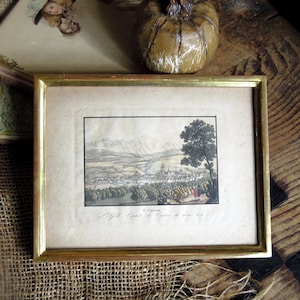 May include: A framed antique print with a gold-colored frame. The print depicts a landscape with a town, mountains, and trees. The print is on aged paper. A decorative pumpkin and other items are in the background.