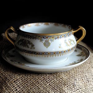 May include: White porcelain soup bowl and saucer set with gold trim and handles. The bowl and saucer feature a decorative floral and geometric pattern in blue, yellow, and gold. The set is placed on a burlap surface.