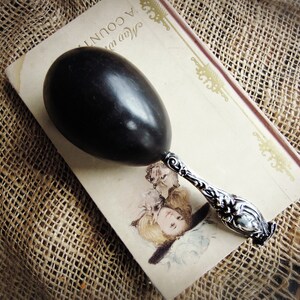 May include: A black egg-shaped rattle with a polished surface and a silver-toned metal handle featuring intricate embossed floral and scrollwork details. The handle is attached to the base of the black oval-shaped percussion instrument.