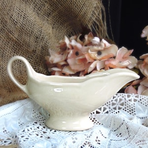 May include: Cream-colored ceramic gravy boat with a handle and a spout. The gravy boat sits on a white lace cloth, with dried flowers in the background. The gravy boat has a decorative pattern on the base.