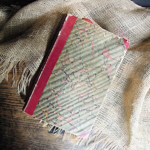 May include: A vintage book with a marbled cover and a red spine. The book is lying on a piece of burlap fabric.