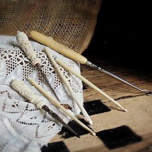 May include: A collection of five vintage needlework tools including two crochet hooks with ornate carved cream-colored handles, two plain cream-colored crochet hooks, and one metal crochet hook with a smooth cream-colored handle.