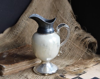 May include: A pitcher featuring a bulbous, pearlescent white glass body, a clear glass handle, and a metallic silver-toned base and pouring spout. The vessel stands on a circular pedestal base and has a curved, elongated spout for pouring liquids.