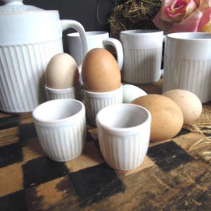 May include: A white ceramic tea set with vertical ribbed detailing, including a teapot, mugs, and egg cups. Several brown and white eggs are displayed in the egg cups and on the surface. The set is arranged on a checkered surface.