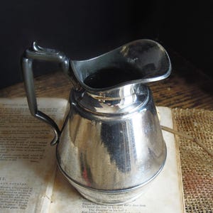 May include: A silver-colored pitcher with a curved handle and a wide spout. The pitcher sits on an open book with aged pages. The background is dark, creating a contrast with the shiny metal.