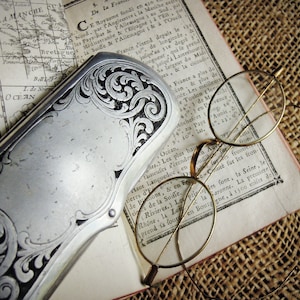 May include: An antique silver-toned vanity case with ornate floral engravings rests on a vintage map and aged paper. Delicate, round-framed eyeglasses with gold-toned wire rims are also present, evoking a sense of history and elegance.