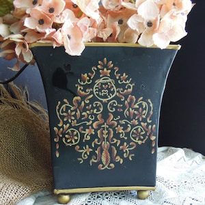 May include: Black decorative vase with gold trim and feet, featuring a detailed floral design in gold and rust colors. The vase is filled with peach-colored artificial flowers, creating a striking contrast.