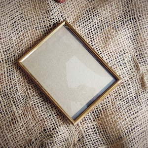 May include: A gold-colored picture frame with a blank interior, sitting on a brown burlap surface.