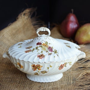 May include: A white porcelain serving dish with a lid, featuring a floral design in shades of yellow, brown, and red. The dish has a scalloped edge and a decorative handle. The dish is on a burlap surface.
