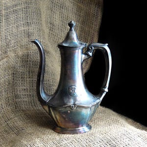 May include: Antique silver-plated coffee pot with a curved spout and ornate handle. The pot features a decorative lid and a detailed emblem on the body. The pot is displayed on a burlap surface.