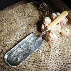 May include: A silver and ivory dustpan with an ornate floral design. The dustpan is lying on a burlap surface with dried roses.