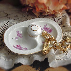 May include: Oval, divided porcelain dish with a white knob and gold trim. The dish features pink floral designs and is accompanied by a gold-toned necklace with faux pearls. The items rest on a lace cloth.